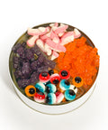 A round tin filled with assorted gummy candies, including pink and white gummy fangs, orange gummy bears, purple gummy bears, and gummy eyeballs in red, blue, white, and orange, on a white background.