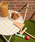 A styled outdoor patio scene with a white round table, a yellow-and-white checkered tablecloth, a glass of iced tea, cookies on a plate, and croquet wands and ball on green artificial turf in front of a brick wall.