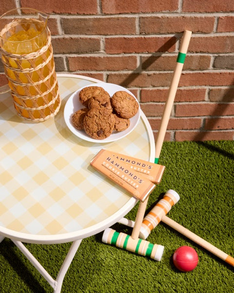 A styled outdoor patio scene with a white round table, a yellow-and-white checkered tablecloth, a glass of iced tea, cookies on a plate, and croquet wands and ball on green artificial turf in front of a brick wall.