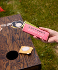 A hand holds a pink and green package of Hammond's Strawberry Matcha in an outdoor setting with a wooden bean bag game on grass.