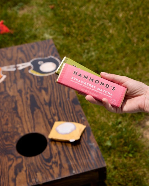 A hand holds a pink and green package of Hammond's Strawberry Matcha in an outdoor setting with a wooden bean bag game on grass.