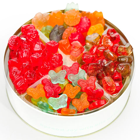 A round white tin filled with assorted colorful gummy candies, including red, orange, green, yellow, and butterfly-shaped gummies, photographed on a white background.