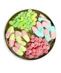 A round metal tin filled with assorted gummy candies arranged in four sections: green sugar-dusted gummies, pale yellow and pink candy slipper gummies, red sugar-dusted gummies, and light blue and pink candy slipper gummies, on a white background.