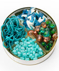 An open round metal tin filled with assorted decorative craft or jewelry components, including bright blue curly strands, turquoise beads, blue-and-white shark-shaped pieces, green and copper shell-shaped beads, and light blue oval beads, all photographed on a white background.